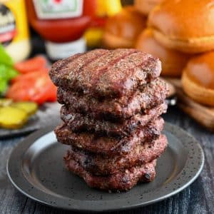 a stack of cooked burgers on a plate with buns, condiments, and fixings behind it
