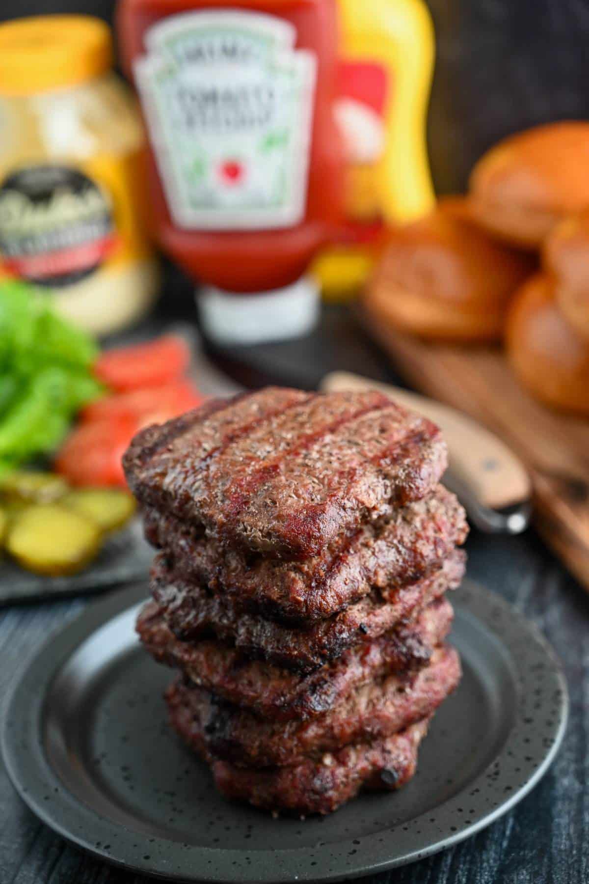 a stack of cooked burgers with condiments and buns in the background