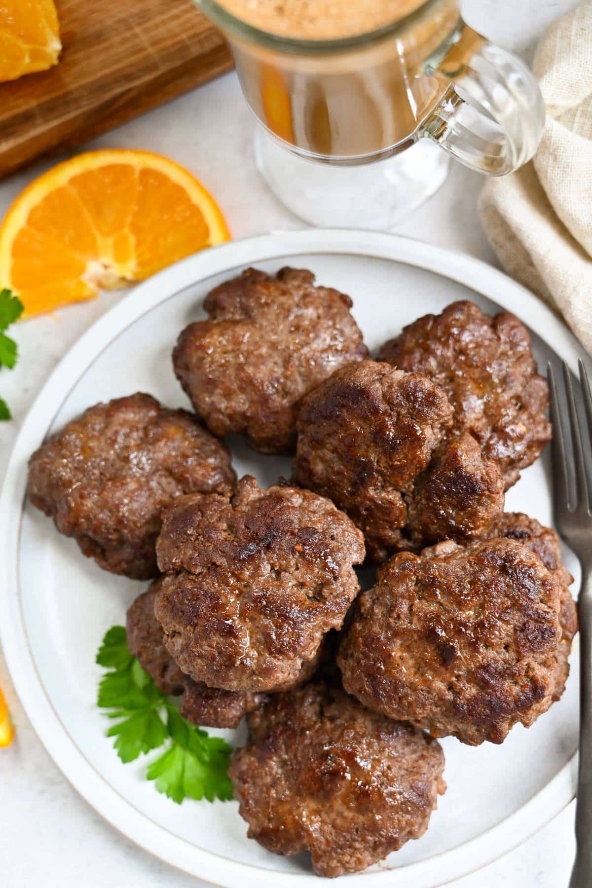 a plate with beef sausage patties and a sprig of parsley with a cup of coffee and orange slices around it