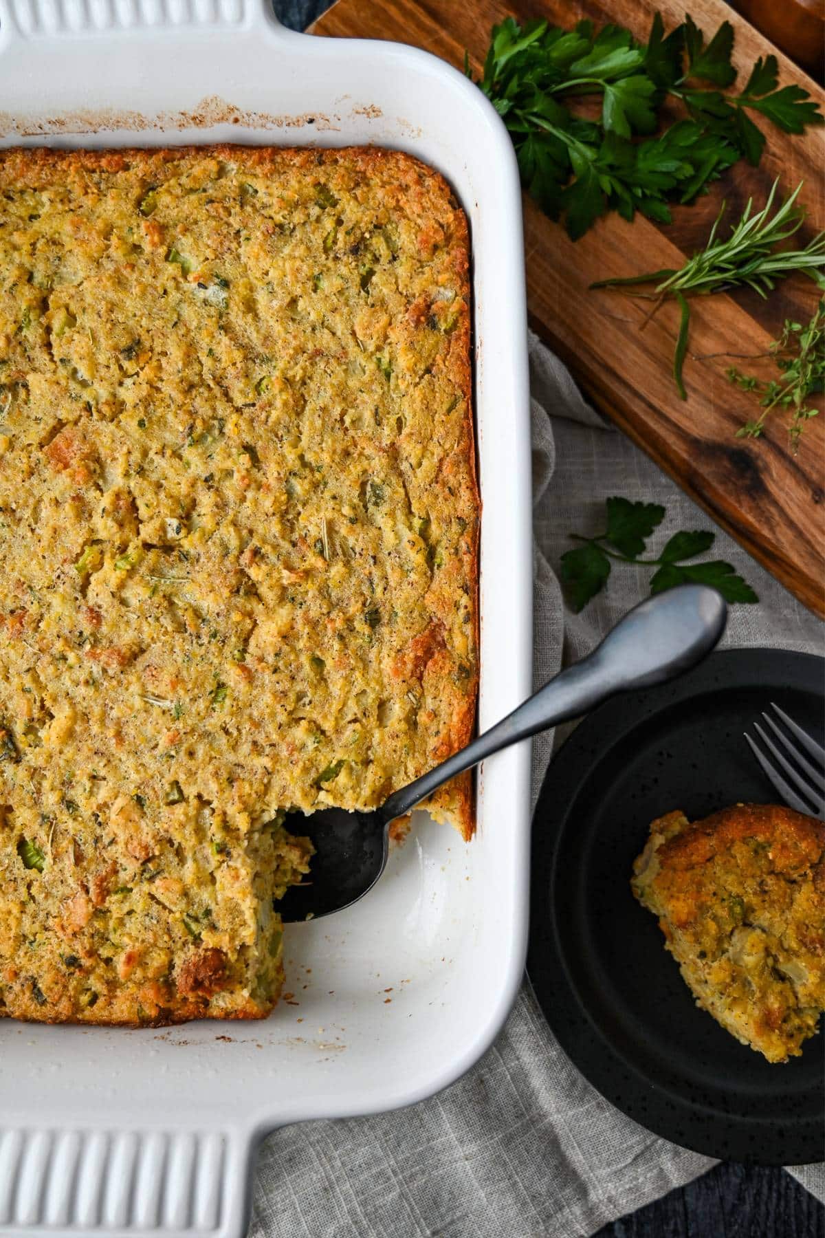 a pan of gluten free cornbread dressing with a spoon and a cutting board with herbs and serving on a plate next to it