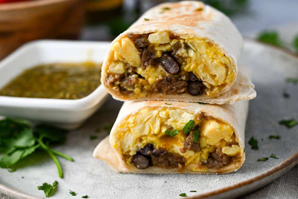 Stacked egg, cheese, and sausage breakfast burritos with green chiles on a plate next to fresh cilantro sprigs and green salsa