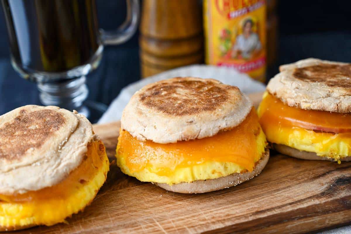 three egg sandwiches with melting cheese on a cutting board with a cup of coffee behind it