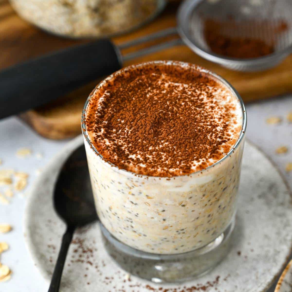tiramisu overnight oats in a glass on a plate with a spoon sprinkled with cocoa powder
