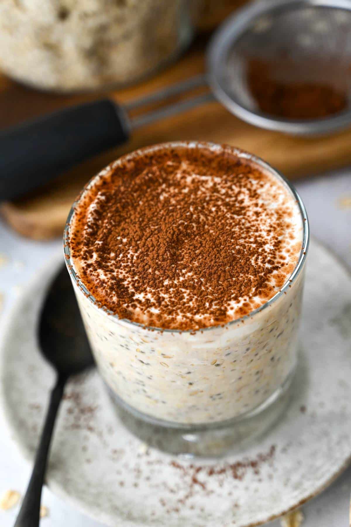 overnight oats in a glass on a plate with a jar of oats and cocoa powder in a strainer behind it