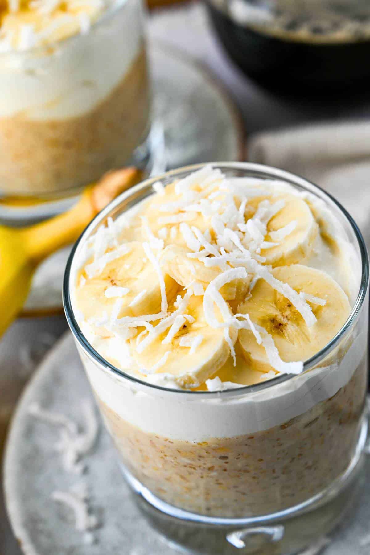 close up of one glass of served banana oats with creamy topping and sliced coconut with another glass of overnight oats behind it and a cup of coffee