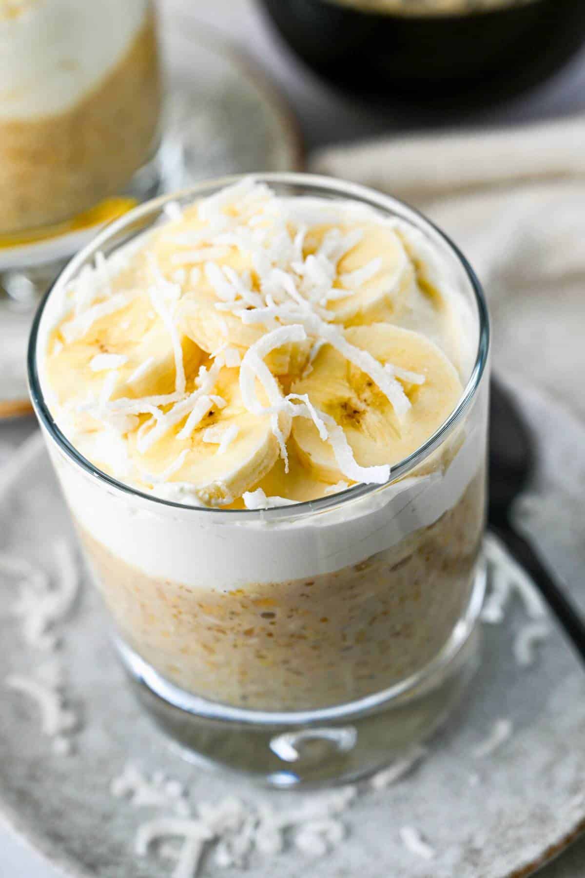 close up of a overnight protein oats topped with a layer of creamy topping, sliced bananas and shredded coconut on a plate with a spoon