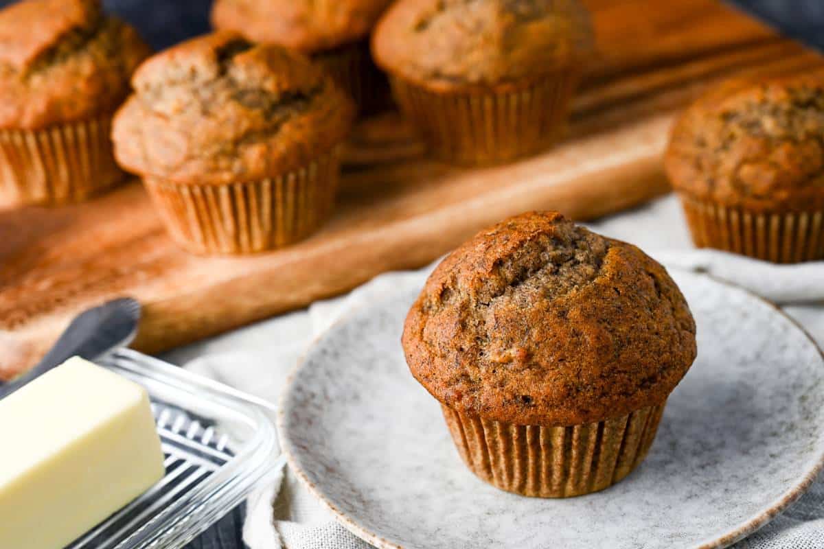 a banana muffin next to a stick of butter in a glass dish with a wooden board with muffins behind it
