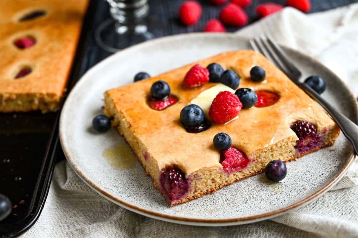 a pan of sheet pan pancakes next to a served slice topped with a pat of butter, syrup and fresh berries
