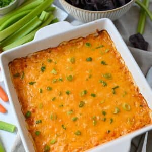 a pan of cheesy baked buffalo chicken dip topped with chopped green onions and served with celery, chips, and carrots