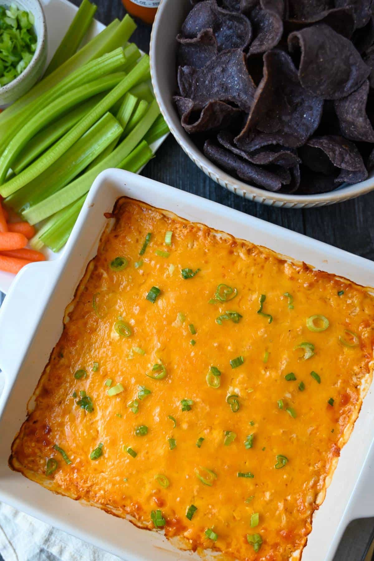 overhead view of a square baking dish of baked buffalo chicken dip with blue corn chips, celery and carrots
