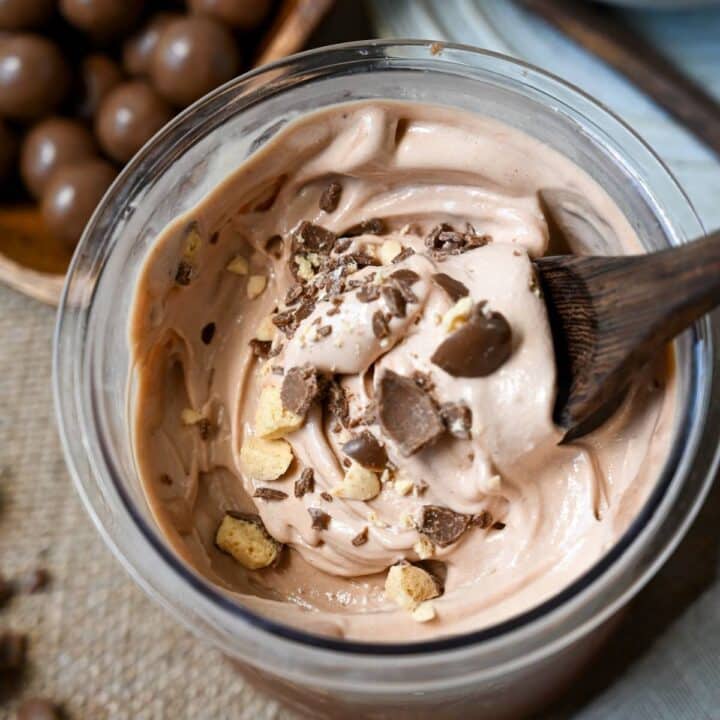 close up of blended chocolate malt ice cream in a ninja creamer jar with crushed malt balls and a spoon with a bowl of malt balls next to it