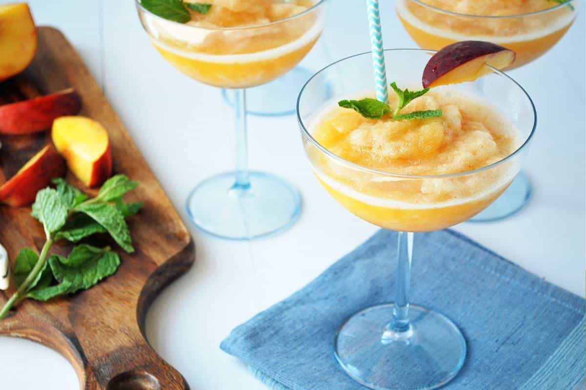 frozen rose with blended peaches in glasses with mint and a peach slice with a cutting board with sliced peaches and mint