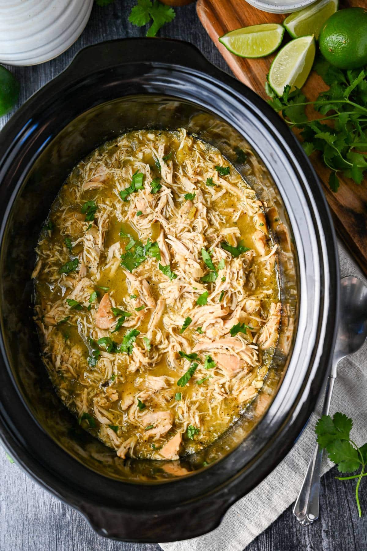 looking down at a crockpot dish with shredded chicken in a green chilli sauce with fresh limes, cilantro and a serving spoon
