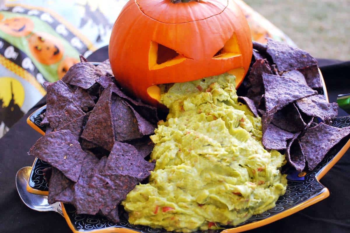 a Halloween themed platter with a carved pie pumpkin with guacamole trailing out of the mouth with blue corn torilla chips