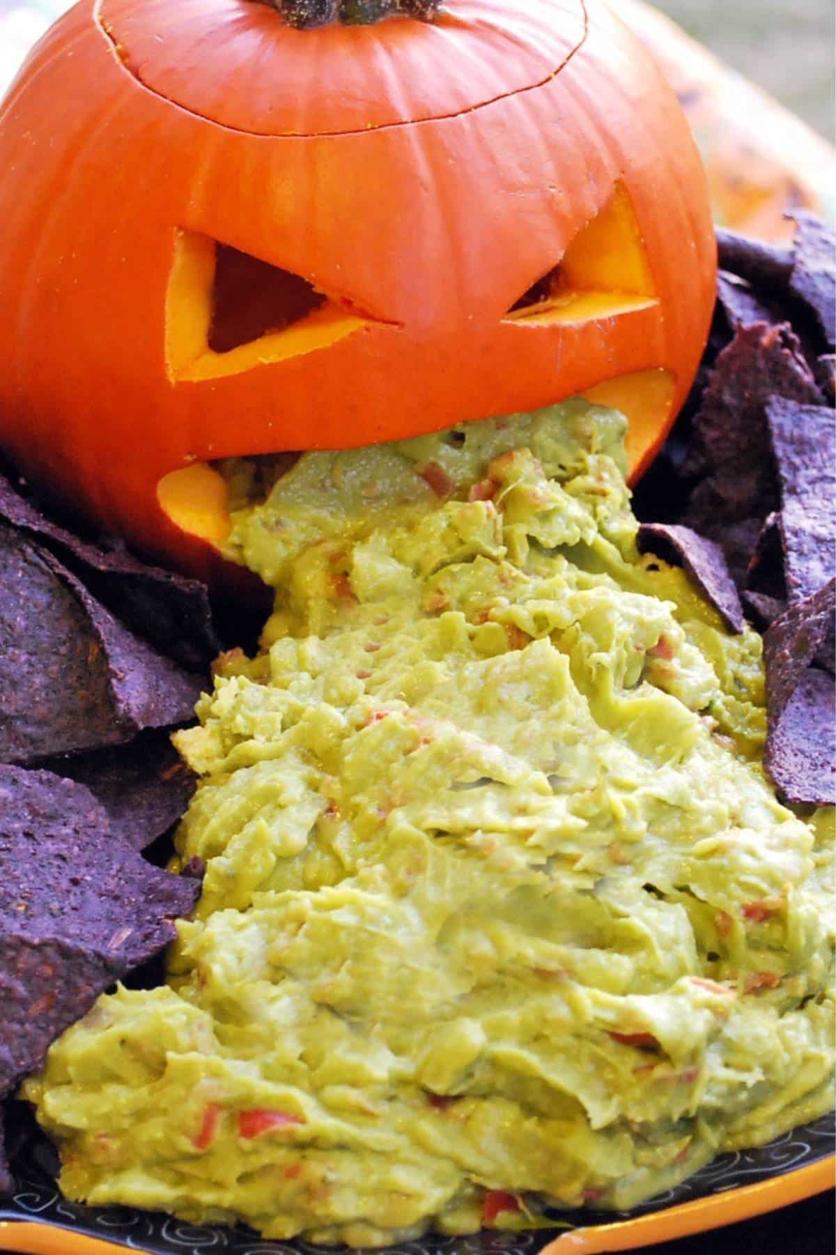 close up of a carved pie pumpkin with guacamole coming out of the mouth with blue corn chips on a platter