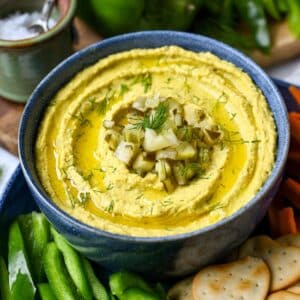 a zoomed in photo of a bowl of dill hummus topped with chopped pickles and sprigs of fresh dill with Naan crackers and sliced peppers