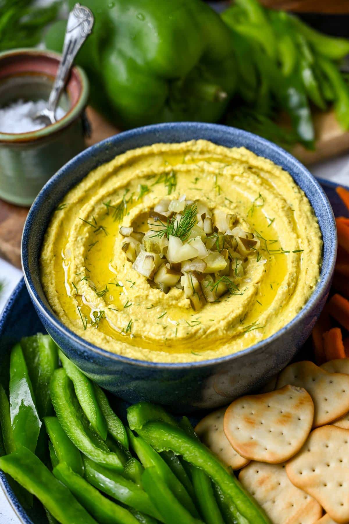 a bowl of dill pickle hummus topped with fresh dill and chopped pickles on a tray with veggies and crackers 