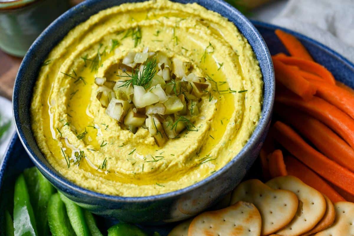 overhead photo of a bowl of hummus with carrot sticks, crackers, sliced peppers and topped with chopped pickles and dill