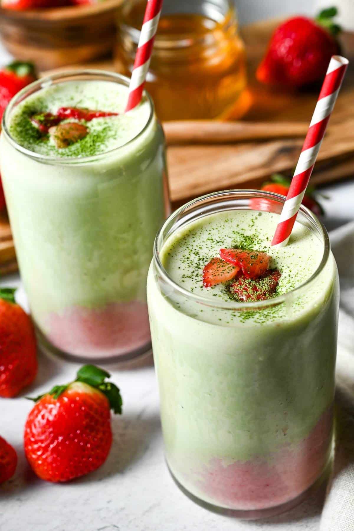 two matcha strawberry layered smoothies in a glass with fresh strawberries and a straw