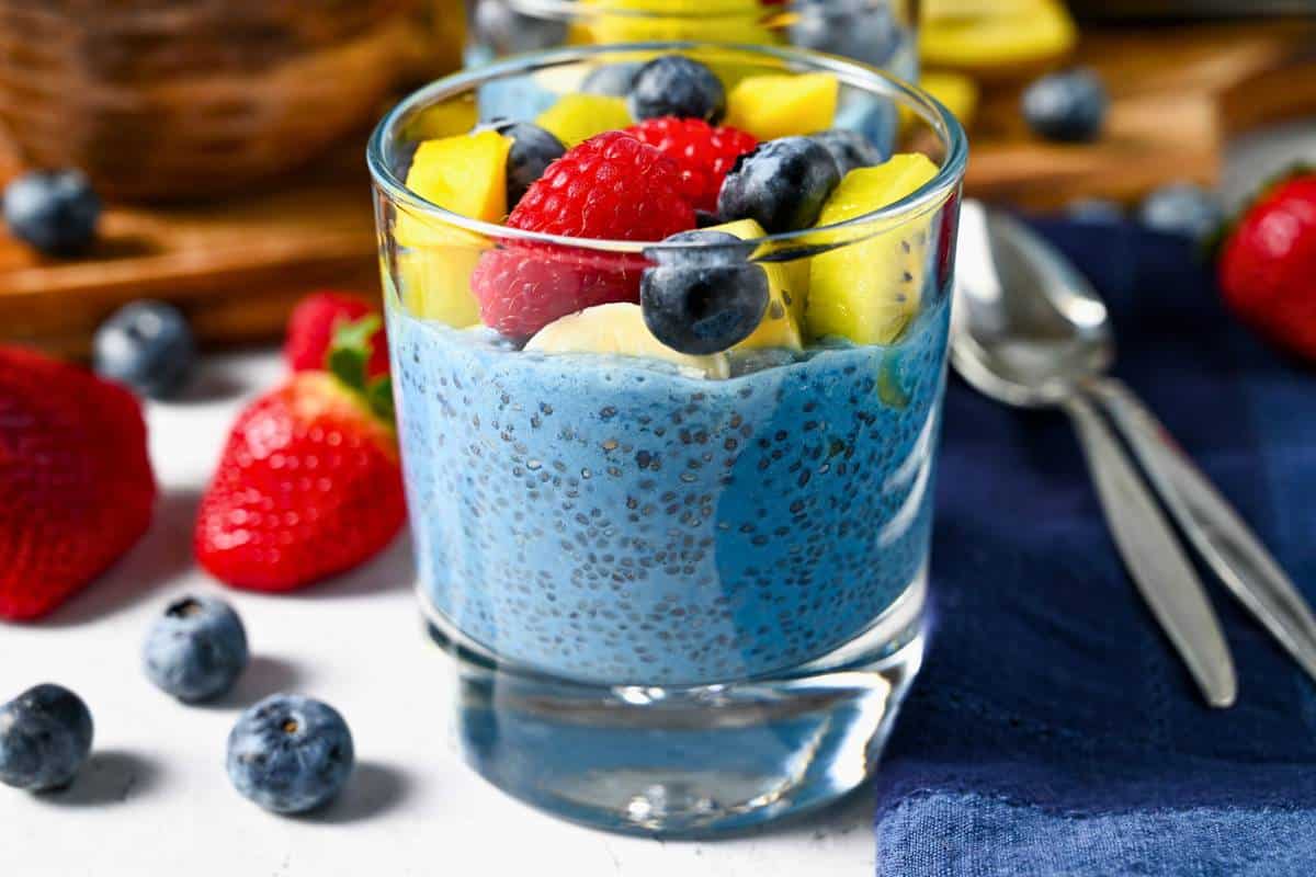 berries on a table surround a glass of blue chia pudding  with a blue napkin and spoons