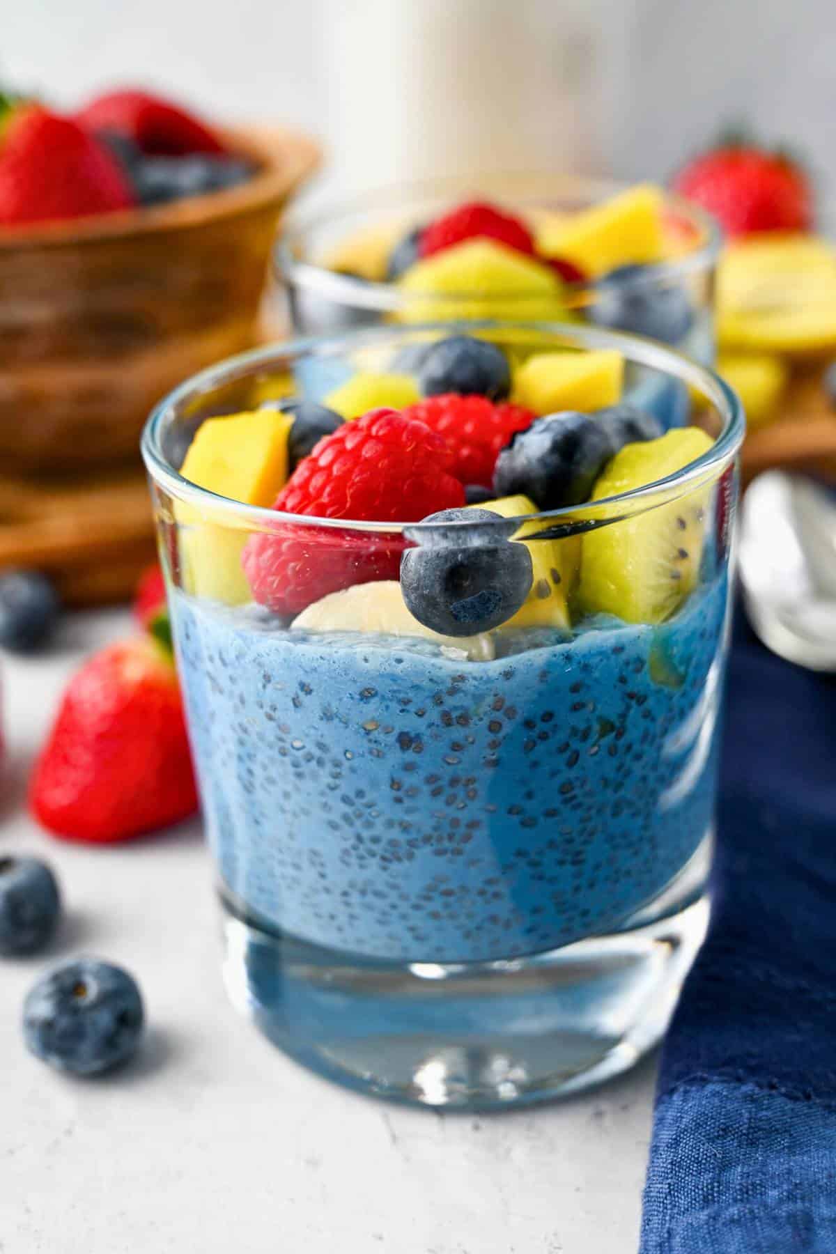 glasses of blue protein chia pudding with berries around them and fruit on top