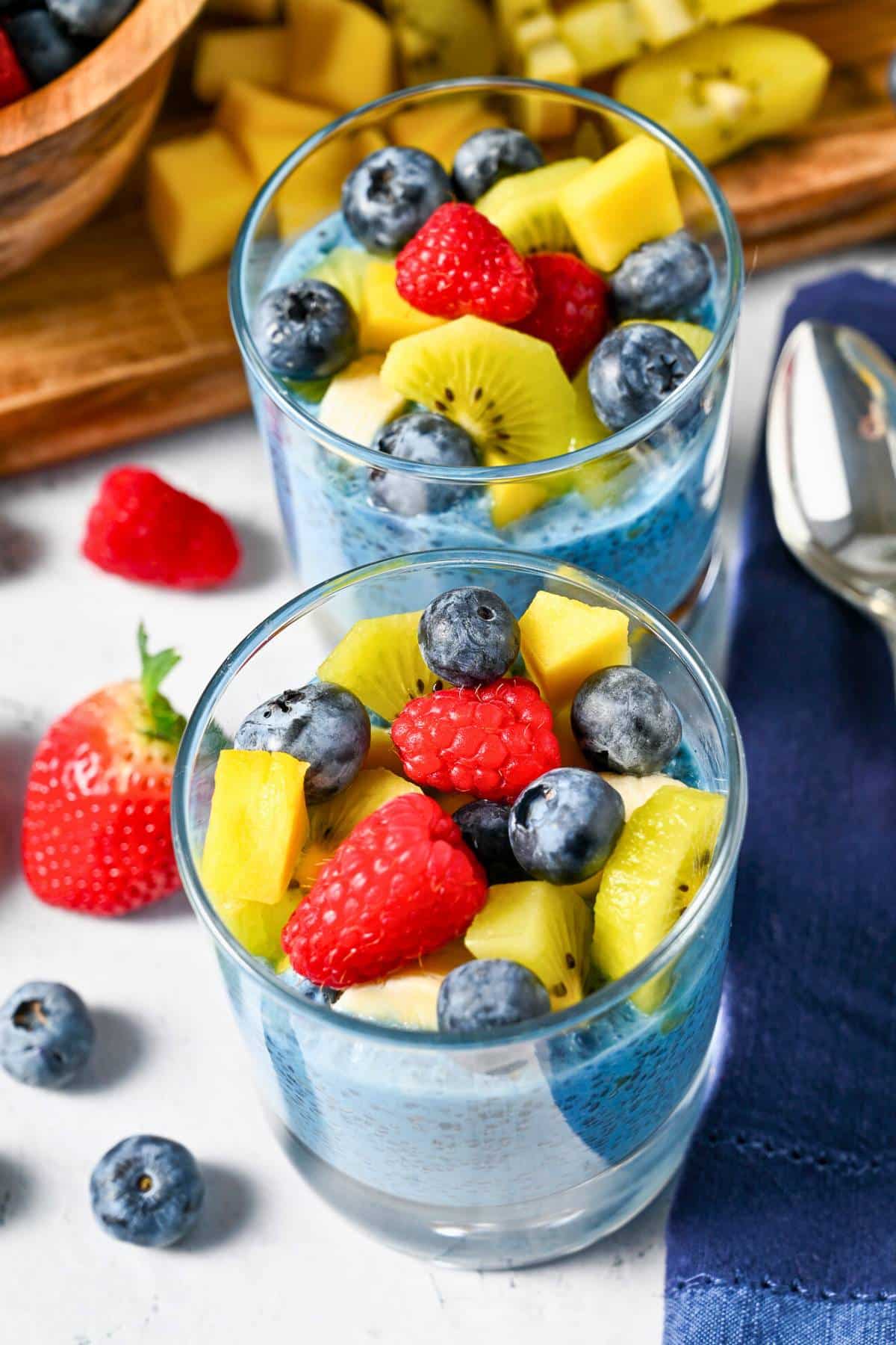 looking down at two chia puddings topped with mixed berries, banana, and mango with a napkin and spoon