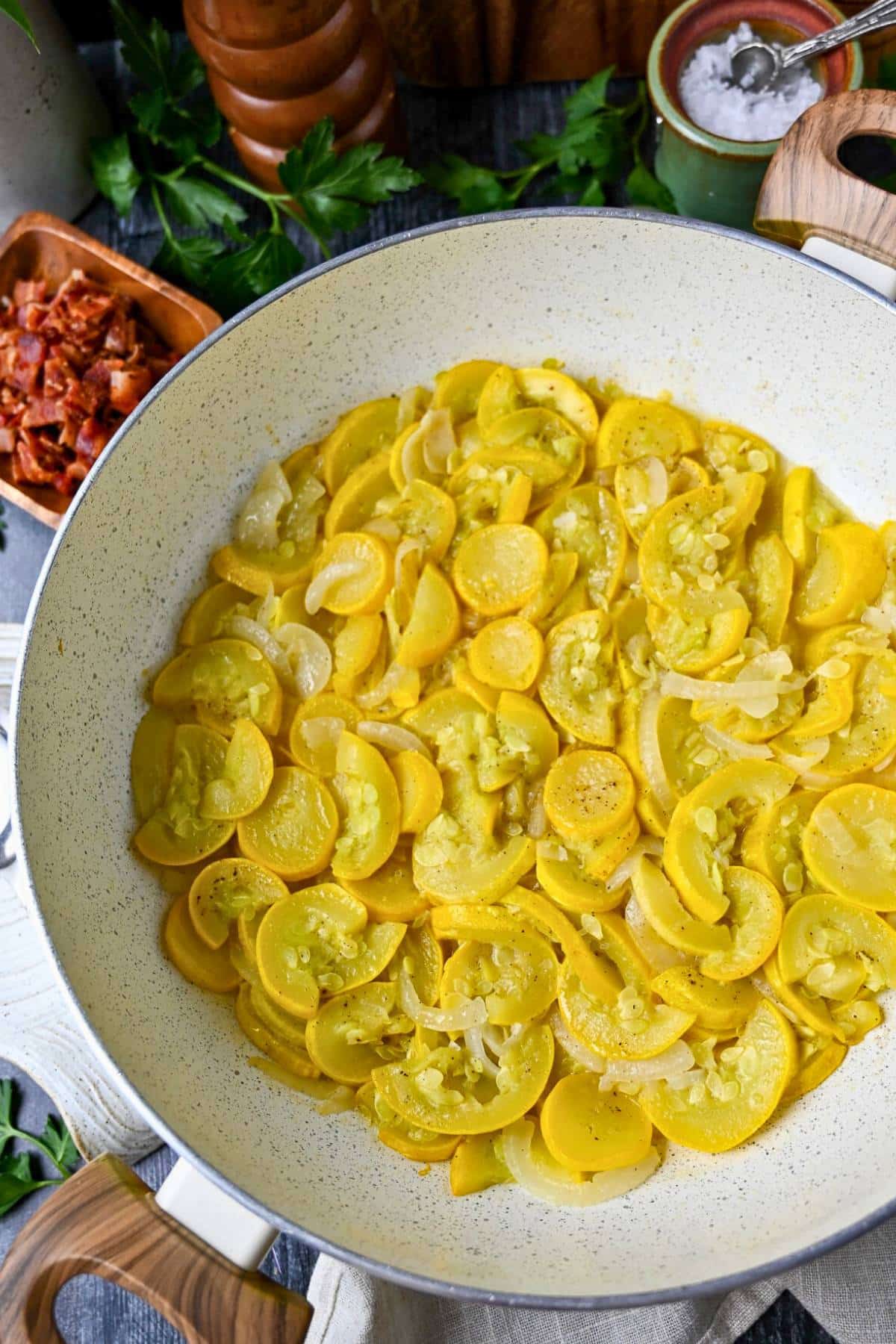 a skillet of stewed squash and onions with a dish of chopped bacon, salt and a pepper mill