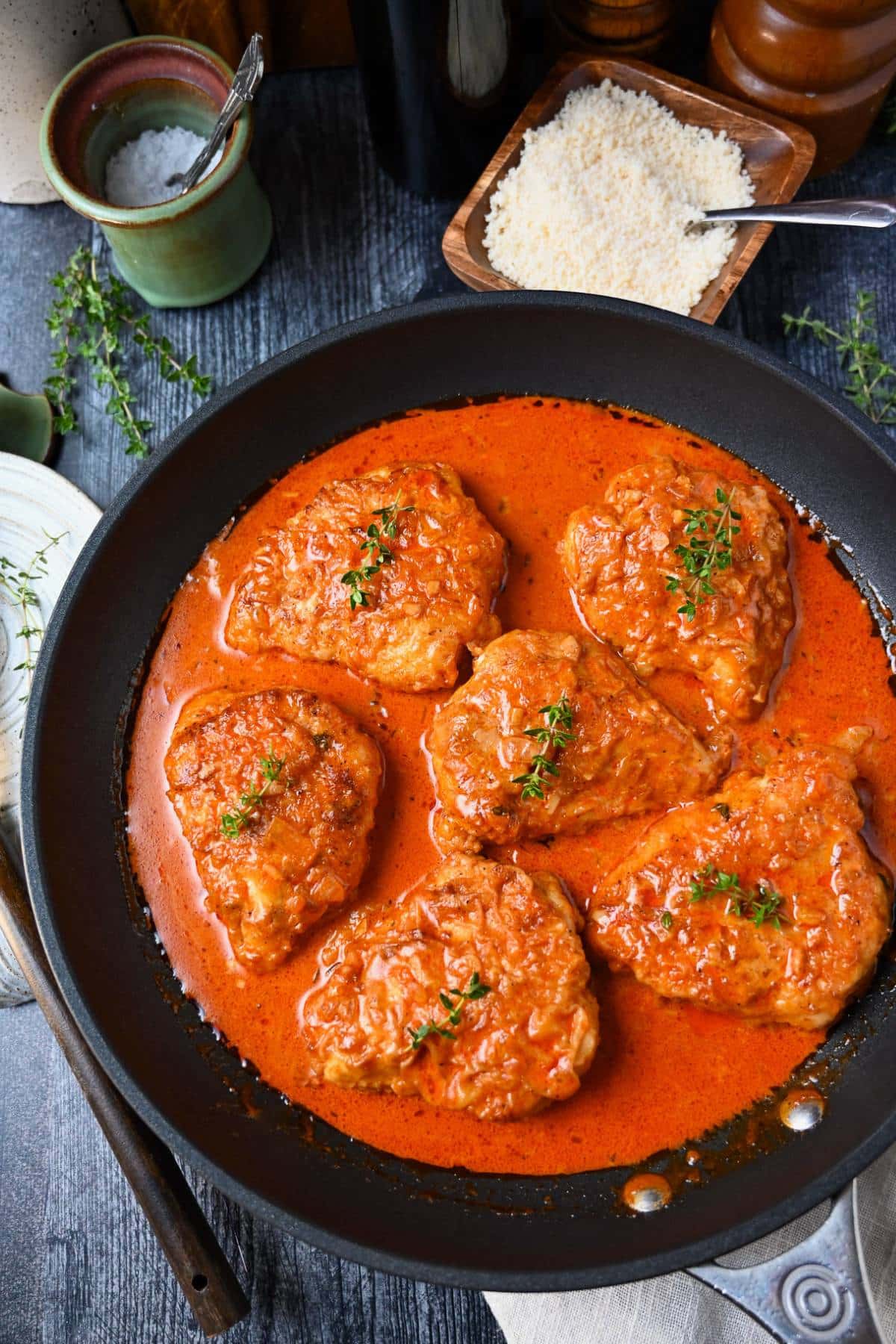 paprika chicken in a skillet with a bowl of parmesan, a dish of salt and fresh thyme sprigs
