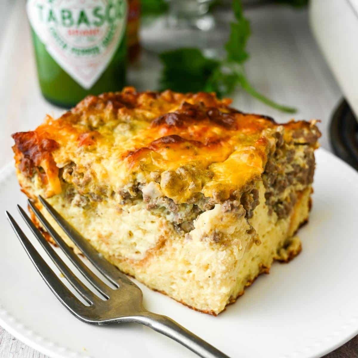 A slice of beef breakfast casserole on a plate with a fork with a bottle of green Tabasco behind it