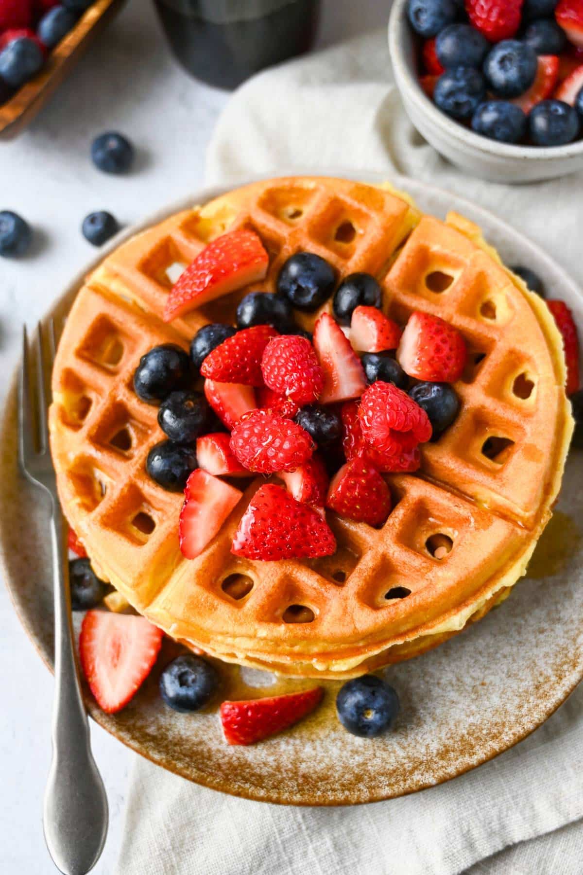 a waffles on a plate with berries with a napkin underneath and a bowl of berries next to it