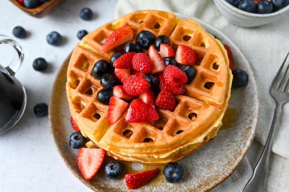 waffles on a plate with fresh fruit and syrup with berries and a fork next to it