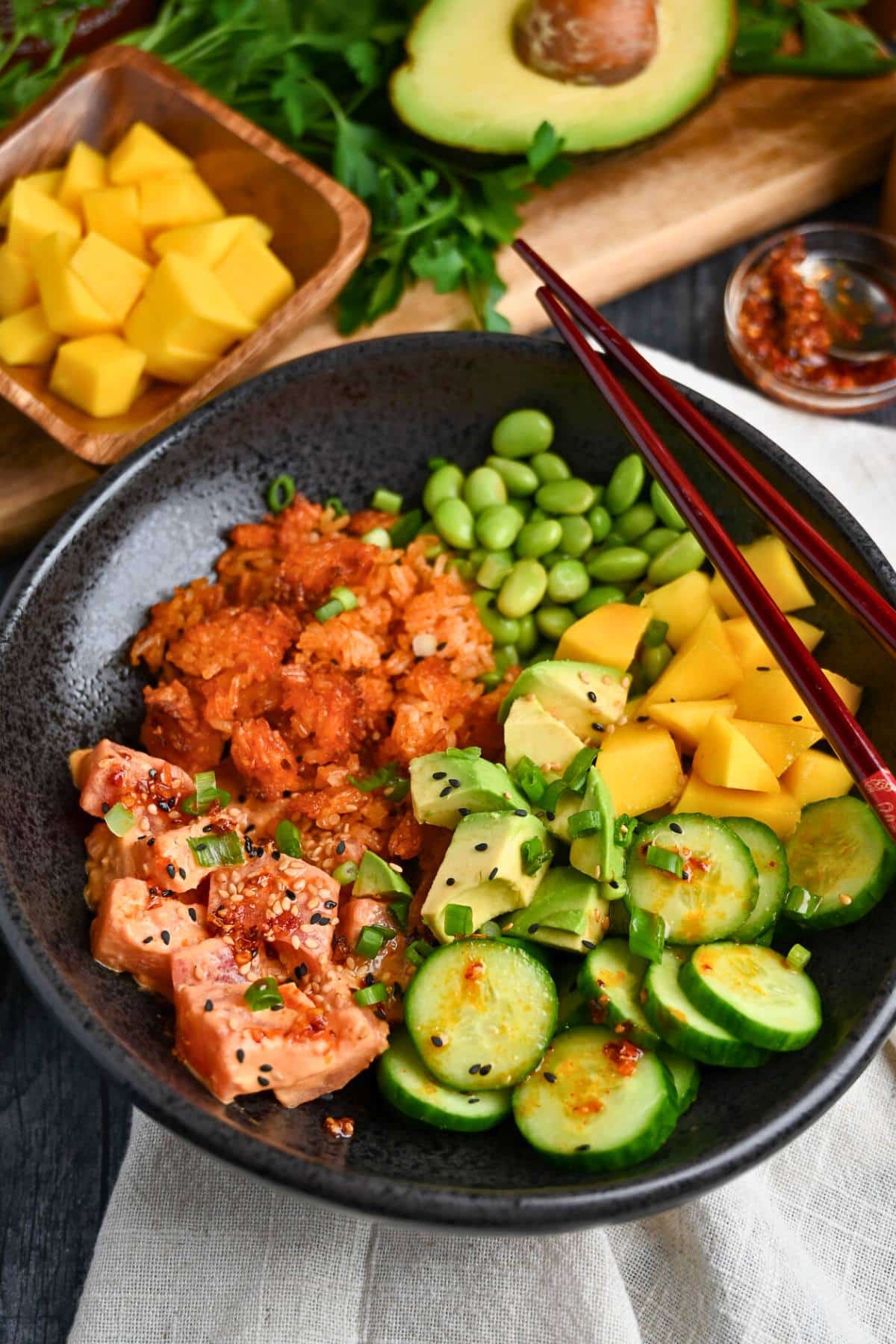 a crispy rice bowl with spicy tuna and veggies with a sliced avocado and a bowl of mango
