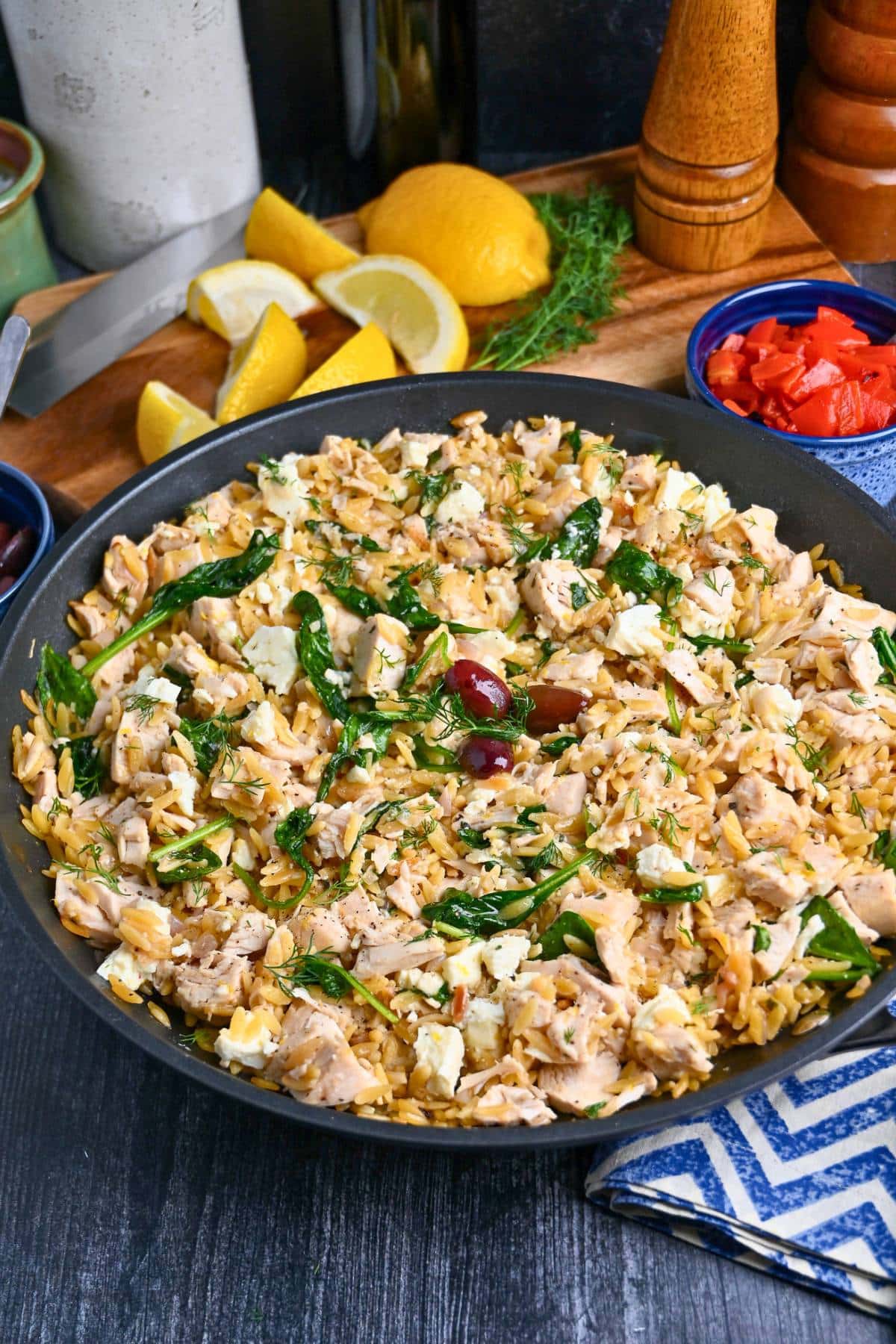 a Greek chicken pasta skillet with sliced lemons and roasted red peppers in the background