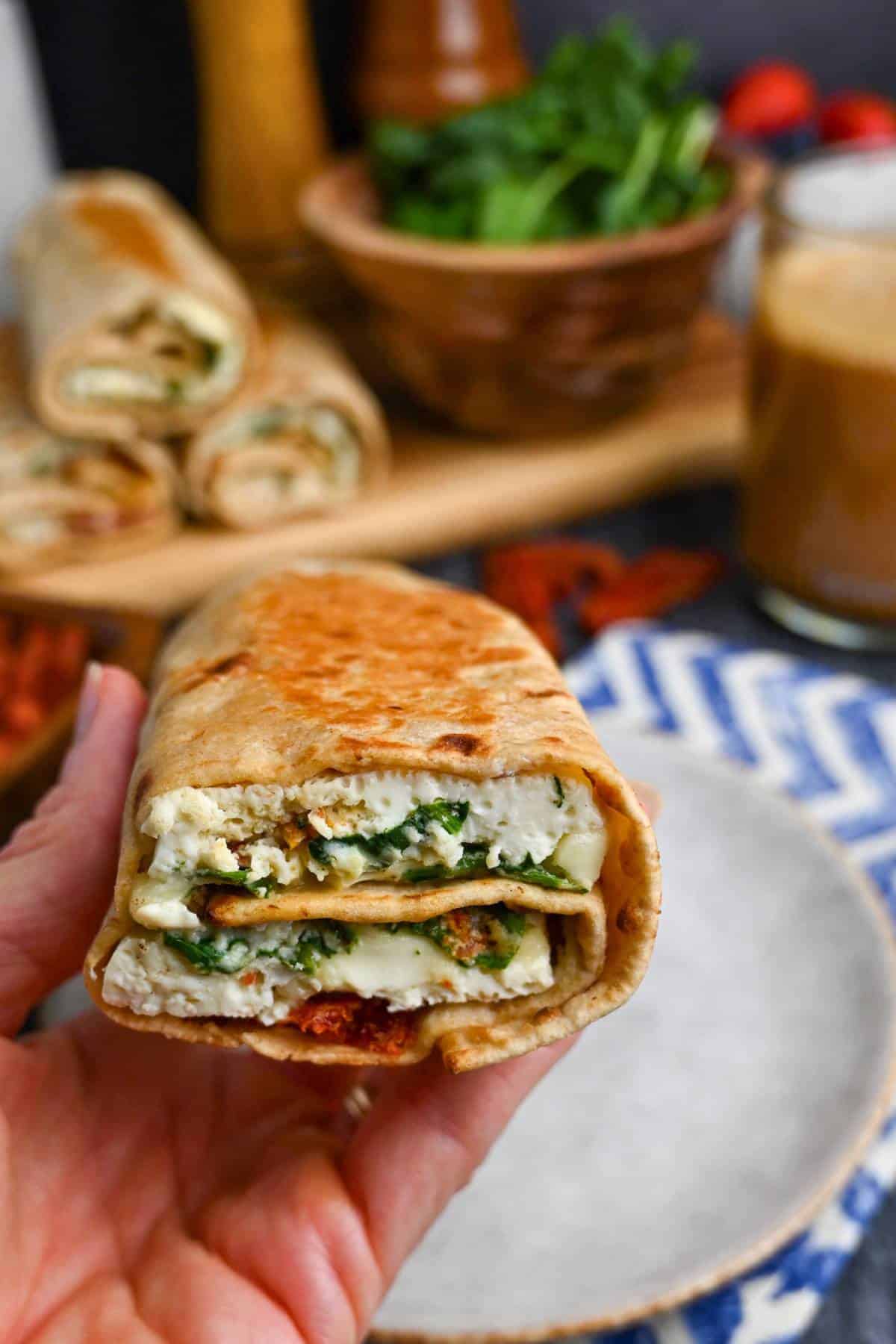 holding up a spinach feta wrap with wraps in the background with a bowl of spinach