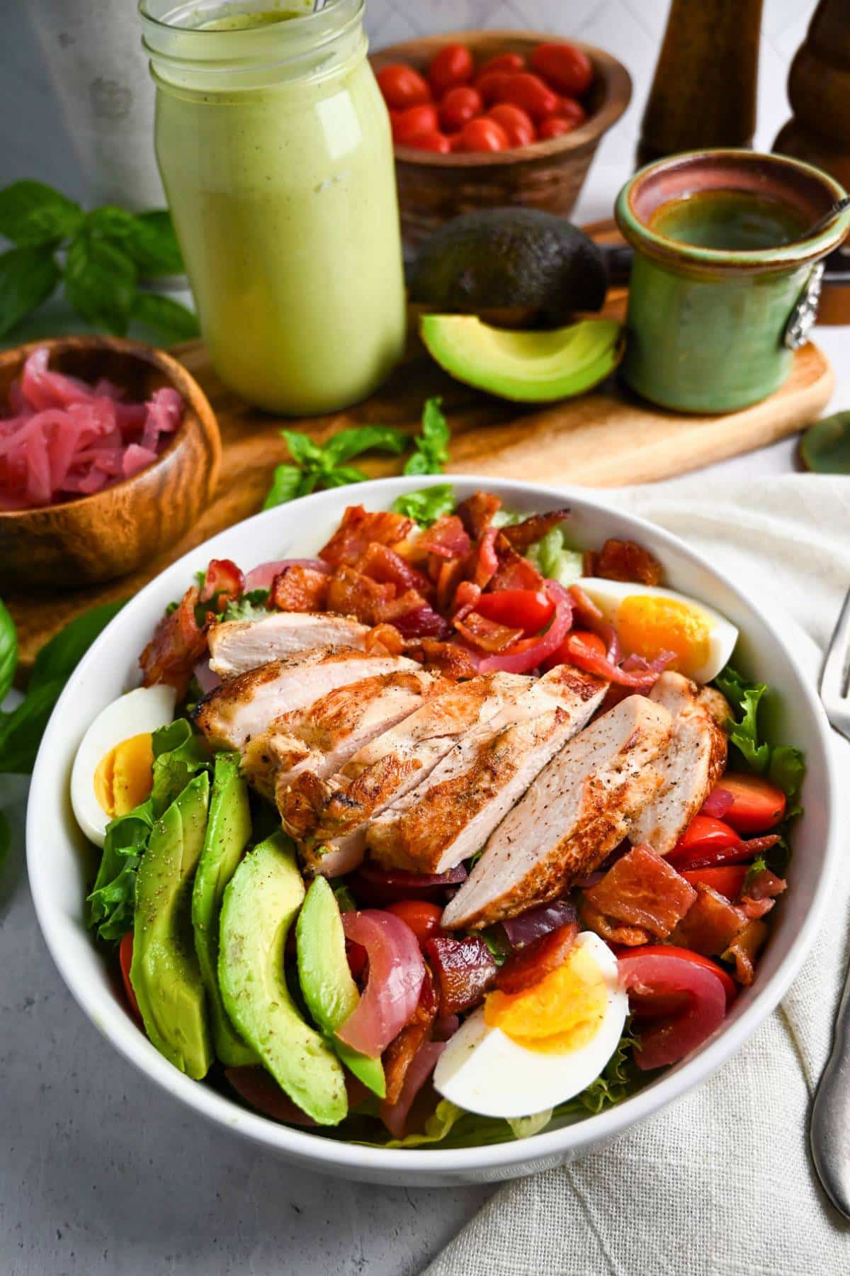 a chicken cobb salad with a jar of green goddess dressing, a bowl of pickled onions, and sliced avocado
